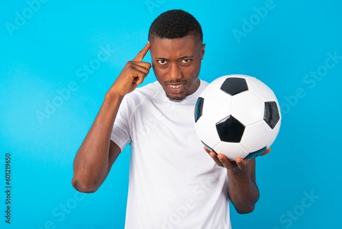 Young man wearing white T-shirt holding a ball over blue background concentrating hard on an idea with a serious look, thinking with both index fingers pointing to forehead.
