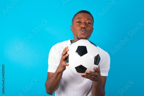 Young man wearing white T-shirt holding a ball over blue background , keeps lips as going to kiss someone, has glad expression, grimace face. Standing indoors. Beauty concept.