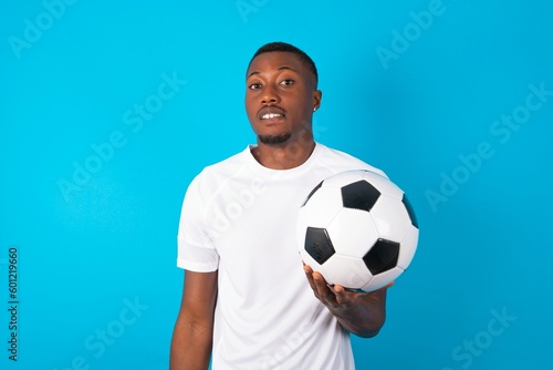 Young man wearing white T-shirt holding a ball over blue background  being nervous and scared biting lips looking camera with impatient expression, pensive. © Roquillo