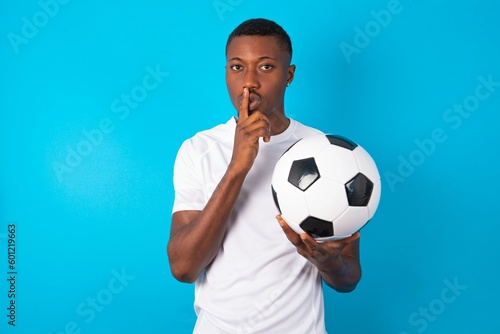 Young man wearing white T-shirt holding a ball over blue background makes hush gesture, asks be quiet. Don't tell my secret or not speak too loud, please!