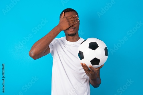 Young man wearing white T-shirt holding a ball over blue background covering eyes with both hands, doesn't want to see anything or feeling ashamed. Human feelings reactions.