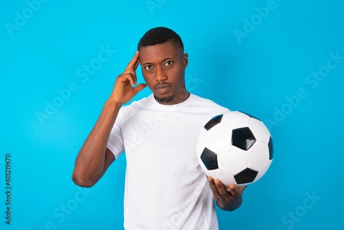 Young man wearing white T-shirt holding a ball over blue background with thoughtful expression, looks away, keeps hand near face, thinks about something pleasant.