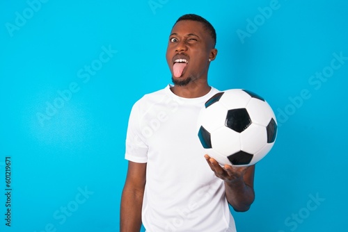Young man wearing white T-shirt holding a ball over blue background with happy and funny face smiling and showing tongue. © Roquillo