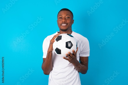 Dreamy charming Young man wearing white T-shirt holding a ball over blue background with pleasant expression, keeps hands crossed near face, excited about something pleasant.