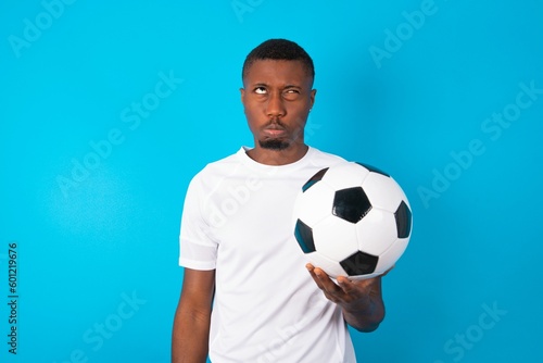 Young man wearing white T-shirt holding a ball over blue background crosses eyes, puts lips, makes grimace with awkward expression has fun alone, plays fool. © Roquillo