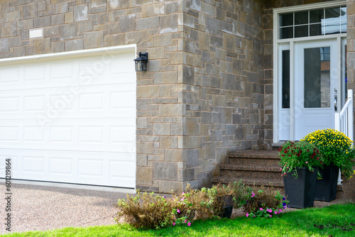 A large white garage door with small glass windows on a beige brick executive house with a white door and steps leading up to a grand entrance. There are multiple shrubs and plants along the steps.
