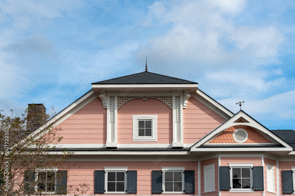 A multi story pink painted wooden house with white trim. The building has multiple small four pane glass windows with black shutters. There's a hip roof with black shingles and a metal weather vane. 
