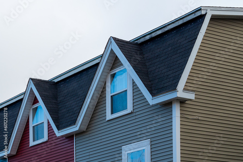 Two adjoined houses with peaked dormers over double hung windows with white trim. The mansard roof has black shingles. The exterior walls are red and tan colored wood siding. The duplex roof is flat. 