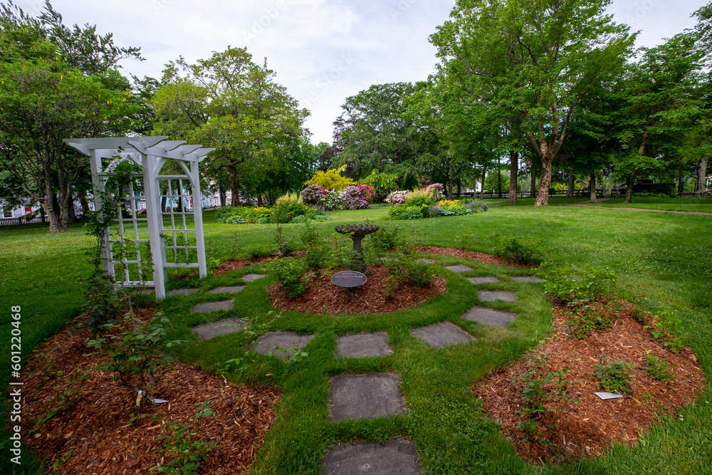 A white wooden archway and square stones forming a circle in a garden with lush tall green maple trees, rose bushes, plants, shrubs  and green grass. The sky is cloudy and blue on a summer's day.