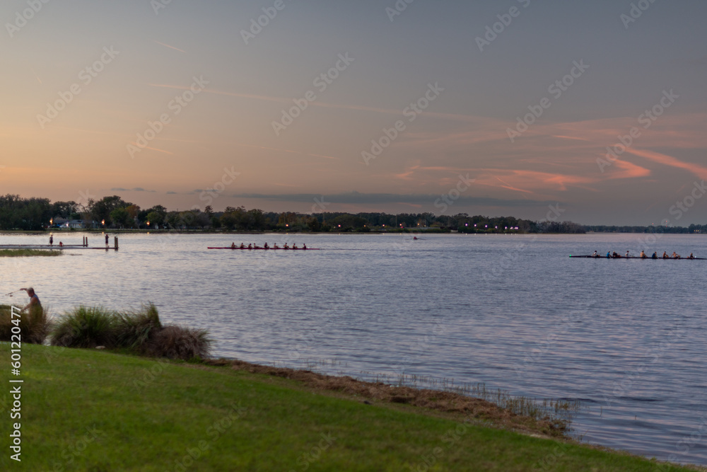 Two teams rowing in shells with multiple teammates at a university competition. The boats are silhouette as the sun sets over the river. There's land with trees in the background. The water is smooth