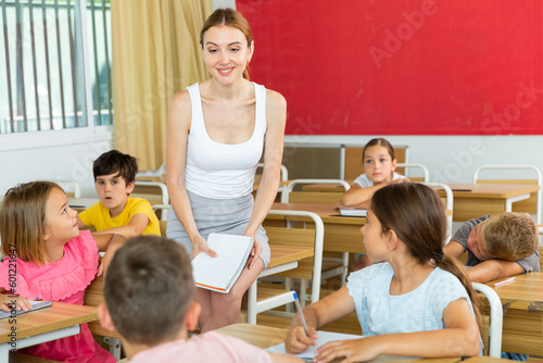 Positive female speaker standing near blackboard and giving lesson for primary school pupils in classroom