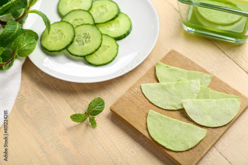 Board with cotton under-eye patches and cucumber slices on light wooden background, closeup
