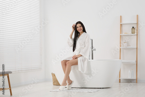 Beautiful happy woman in stylish bathrobe sitting on tub in bathroom