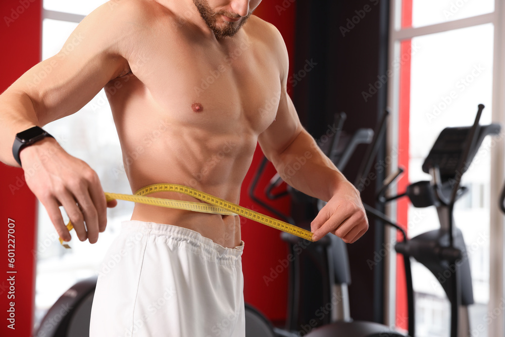 Naklejka premium Athletic man measuring waist with tape in gym, closeup