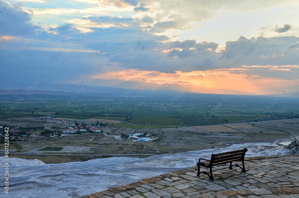 An empty bench in Pamukkale, Turkey, facing the immensity of the horizon, houses, hotels, green fields, and a sunset coloring the blue sky with some clouds in orange.