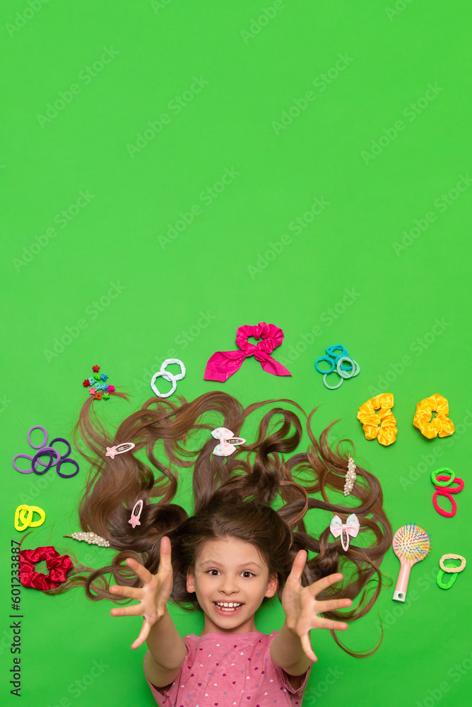 A little girl with long hair lies with elastic bands and hair clips and accessories. Hairstyles for girls. Green isolated background. Copy space. Space for text.