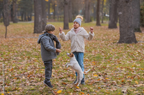 Caucasian children are walking with jack russell terrier in autumn park. Boy, girl and dog are jumping outdoors.