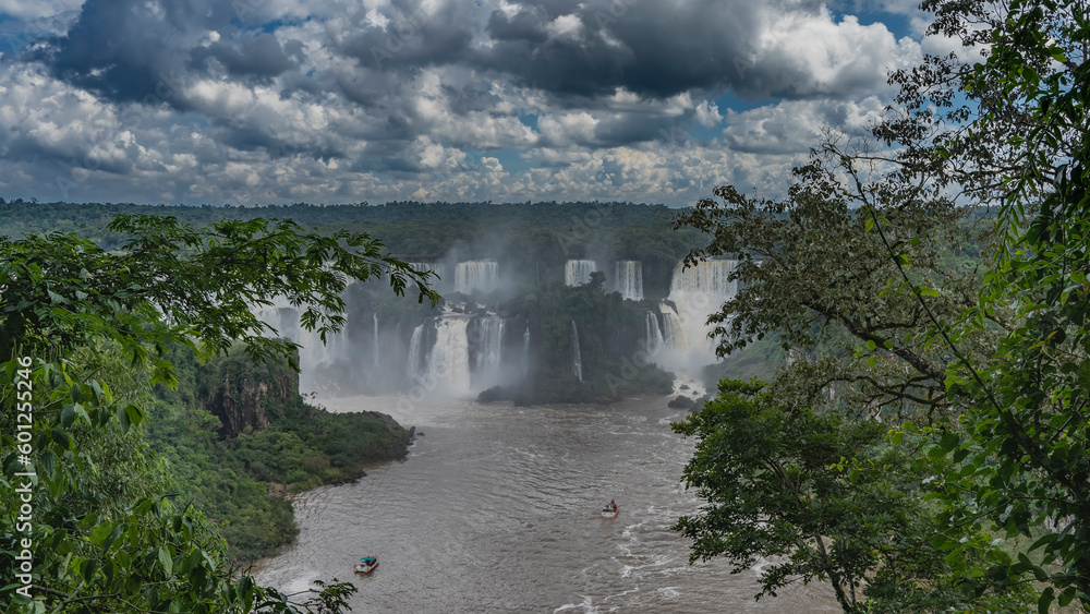 Streams of water cascades down into the river. Tourist boats are visible in the riverbed. Fog in the air. Tropical vegetation around. Clouds in the sky. Iguazu Falls. Brazil.