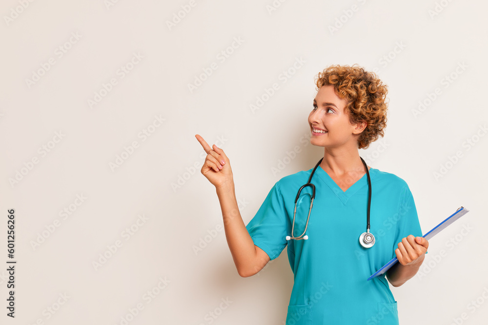 Young professional medical worker in turquoise uniform and with stethoscope poses aside on white background, smiling and pointing finger on the right, good doctor concept, copy space Young professional medical worker in turquoise uniform and with stethoscope poses aside on white background, smiling and pointing finger on the right, good doctor concept, copy space