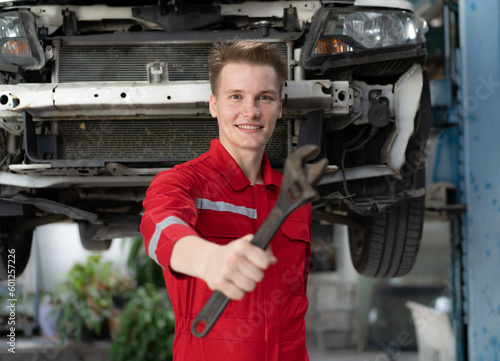 Auto mechanic in red uniform standing at car repair station holding wrench prepare to fix broken engine vehicles at mechanics garage. Smiling maintenance engineer happy work in automotive industry