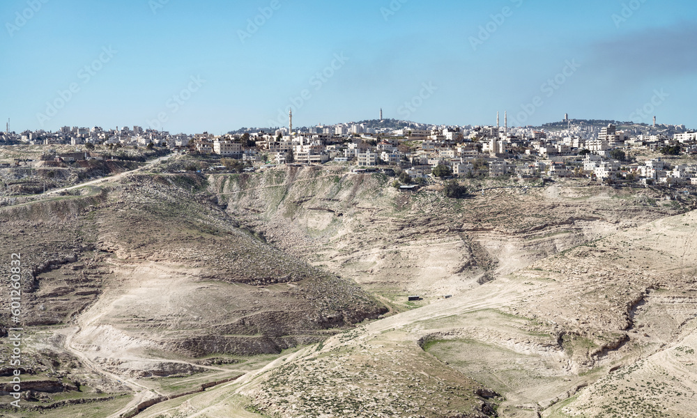 the ancient Palestinian town of al-Azariya sits in front of the towers of East Jerusalem and behind barren desert hills with a blue sky in the background