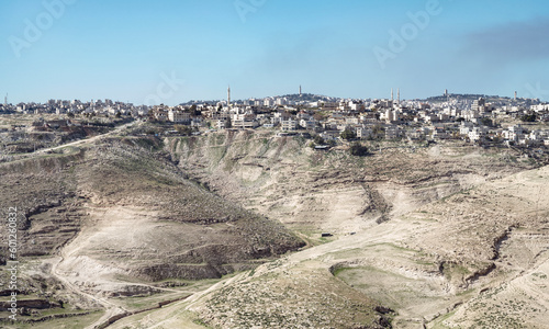 the ancient Palestinian town of al-Azariya sits in front of the towers of East Jerusalem and behind barren desert hills with a blue sky in the background