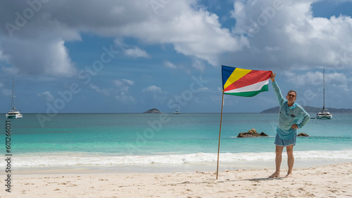 A man stands on the white sand of the beach next to the flag of the Seychelles  holding it by the corner. Yachts are visible in the turquoise ocean. Silhouettes of islands on the horizon. Praslin. 