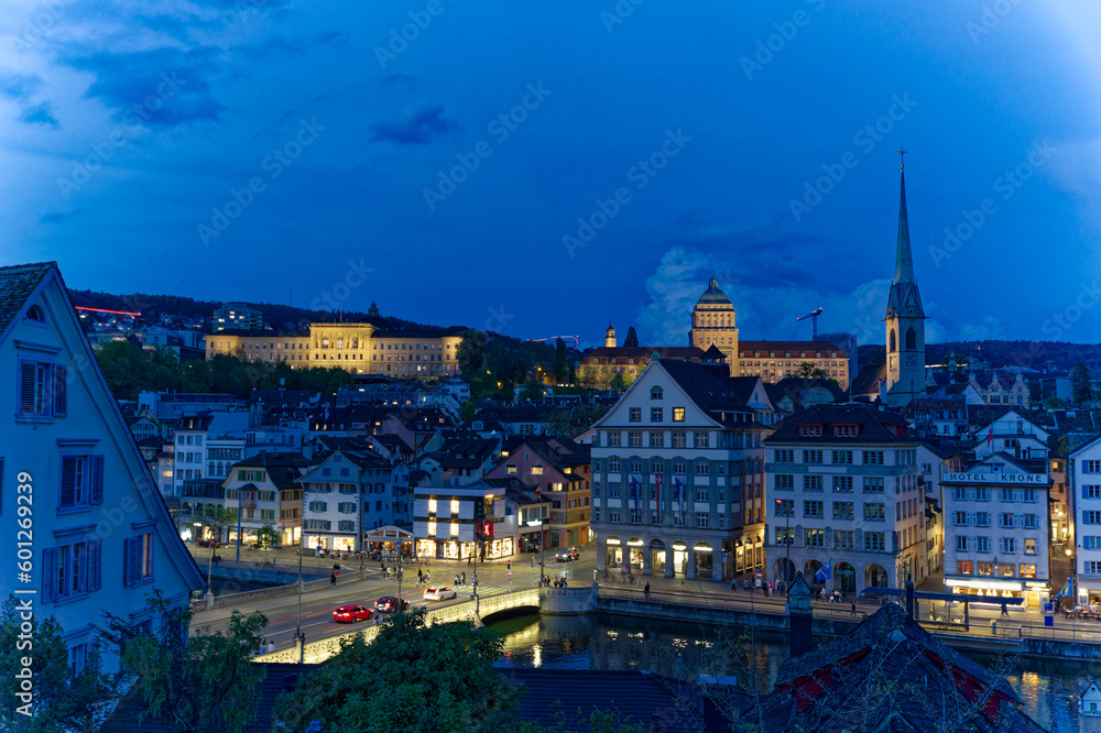 Fototapeta premium Aerial view over the old town of Zürich with Limmat River and city lights on a spring evening with dramatic sky. Photo taken May 6th, 2023, Zurich, Switzerland.