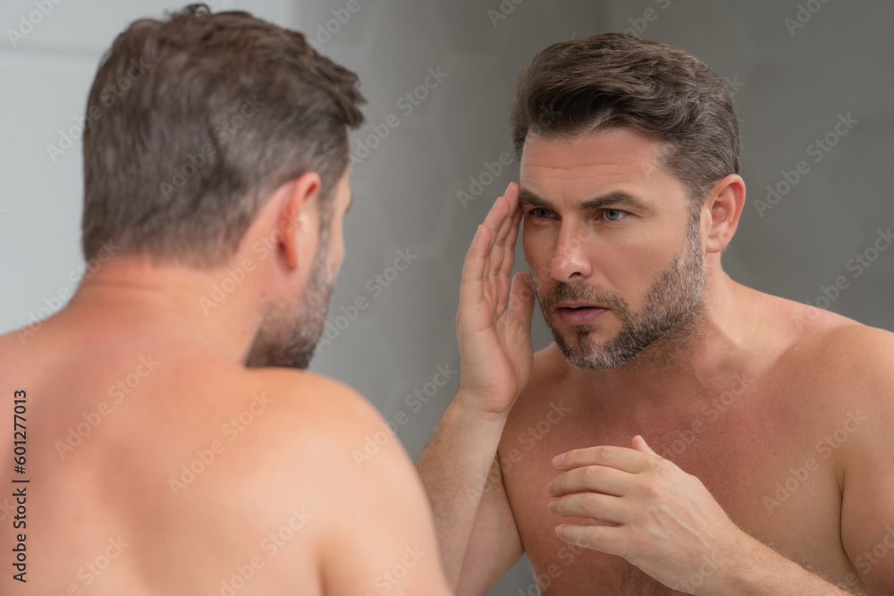 Man applying cosmetic face cream on mirror. Facial treatment. Portrait of man with bare naked shoulders touching skin. Sexy man with skin care product. Male face creme. Perfect skin, morning routine. Man applying cosmetic face cream on mirror. Facial treatment. Portrait of man with bare naked shoulders touching skin. Sexy man with skin care product. Male face creme. Perfect skin, morning routine.