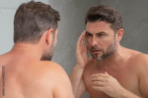 Man applying cosmetic face cream on mirror. Facial treatment. Portrait of man with bare naked shoulders touching skin. Sexy man with skin care product. Male face creme. Perfect skin, morning routine.