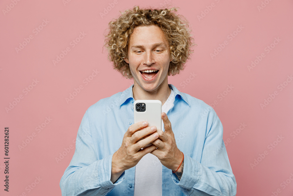 Young smiling happy fun cheerful caucasian blond man wear blue shirt white t-shirt hold in hand use mobile cell phone isolated on plain pastel light pink background studio portrait. Lifestyle concept.
