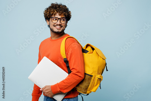 Sideways oung teen IT Indian boy student wear casual clothes backpack bag work hold use laptop pc computer look aside isolated on plain pastel blue background. High school university college concept.