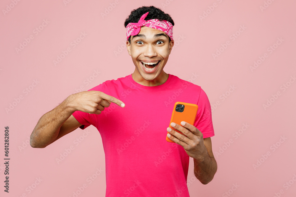 Young man of African American ethnicity wear headscarf t-shirt casual clothes hold in hand use point finger on mobile cell phone isolated on pastel plain light pink color background studio portrait. Young man of African American ethnicity wear headscarf t-shirt casual clothes hold in hand use point finger on mobile cell phone isolated on pastel plain light pink color background studio portrait.