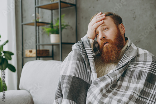 Young sad ginger man wrapped in plaid holding head sits on grey sofa couch stay at home hotel flat spend time in living room indoor. Healthy lifestyle ill sick disease treatment cold season concept. photo