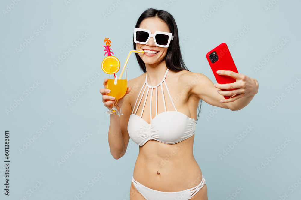 Young woman wear swimsuit glasses hold orange juice cocktail do selfie shot mobile cell phone near hotel pool isolated on plain pastel light blue background. Summer vacation sea rest sun tan concept. Young woman wear swimsuit glasses hold orange juice cocktail do selfie shot mobile cell phone near hotel pool isolated on plain pastel light blue background. Summer vacation sea rest sun tan concept.