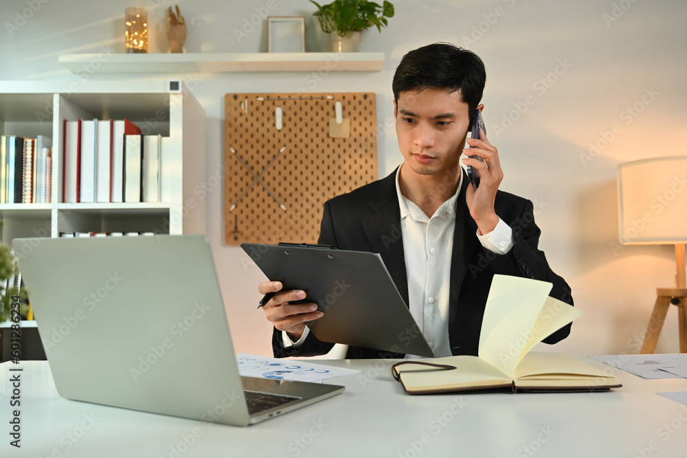 Focused male financial advisor consulting client distantly by mobile phone call, and using laptop at workplace Focused male financial advisor consulting client distantly by mobile phone call, and using laptop at workplace