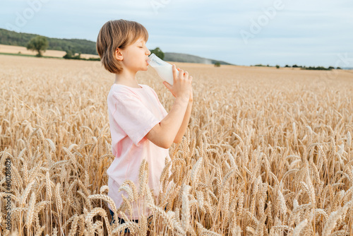 Cute happy girl drinks fresh milk from a glass bottle while standing in a field of cereal spikelets. The concept of healthy food and bio organic products.