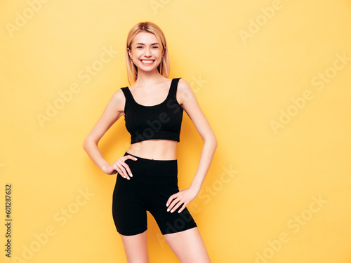 Young beautiful smiling blond female in summer black cycling shorts costume clothes. Sexy carefree woman posing near yellow wall in studio. Positive model having fun indoors. Cheerful and happy