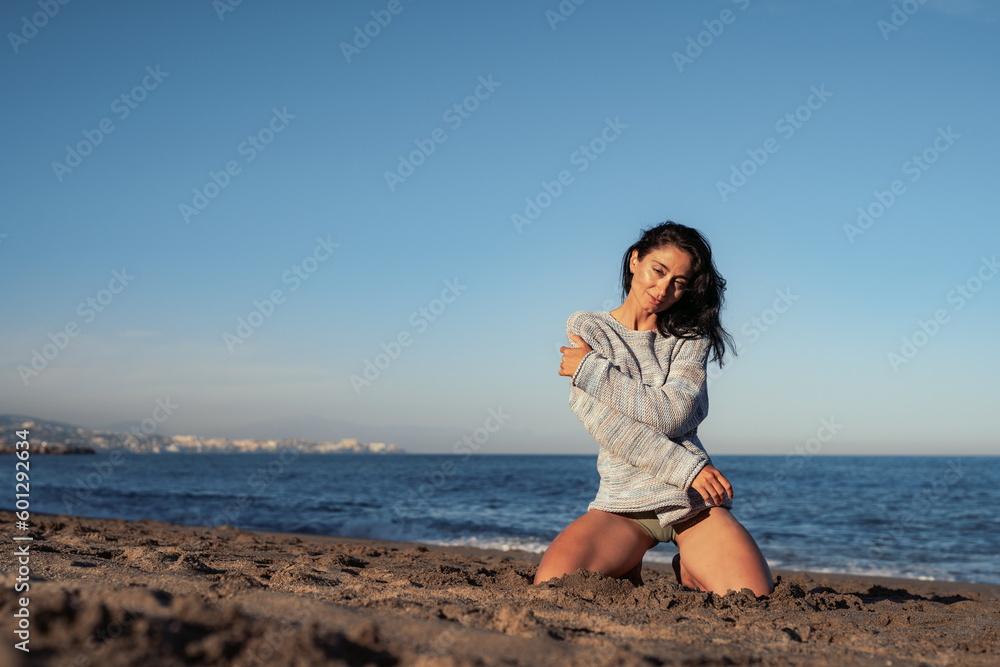 beautiful brunette woman in a sweater and swimsuit sits on the sand by the sea enjoys the environment