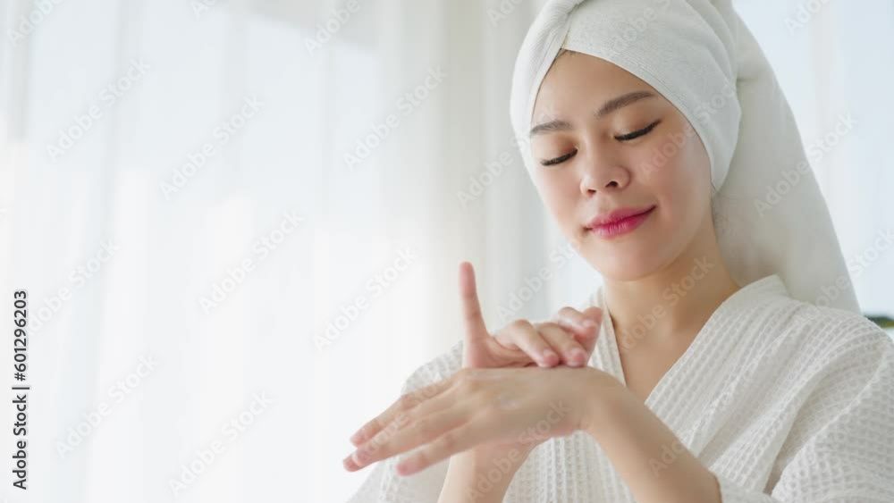 Beautiful Asian woman in white robe and towel on head applying moisturizing cream on hand for protection skin after shower in the morning at bedroom. Beauty and body care concept Beautiful Asian woman in white robe and towel on head applying moisturizing cream on hand for protection skin after shower in the morning at bedroom. Beauty and body care concept