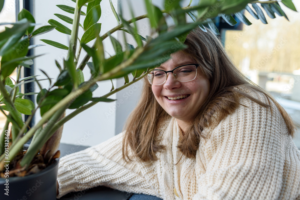 Smiling young woman in knitted sweater sitting near window with home plant Smiling young woman in knitted sweater sitting near window with home plant
