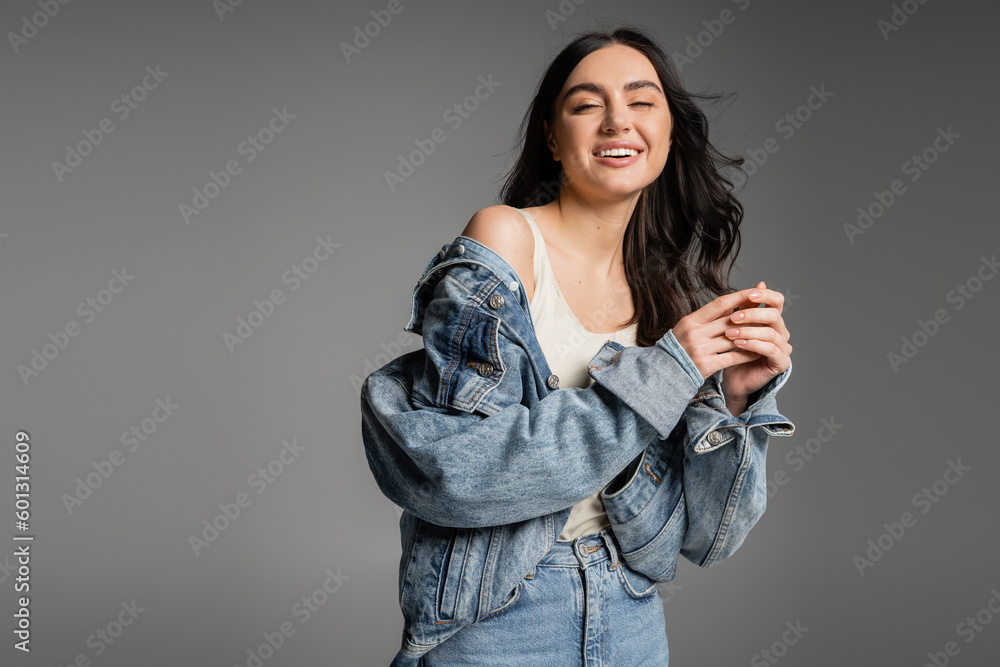 positive young woman with brunette hair standing in blue jeans and fashionable denim jacket while smiling and posing isolated on grey background positive young woman with brunette hair standing in blue jeans and fashionable denim jacket while smiling and posing isolated on grey background