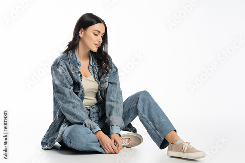 full length of alluring young woman with natural makeup and brunette hair posing in trendy denim outfit while sitting and looking away on white background