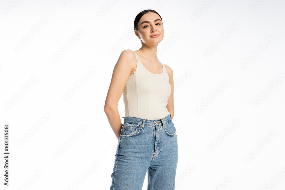 appealing and young woman with natural makeup, brunette hair and perfect skin smiling while posing in tank top and denim jeans and looking at camera on white background appealing and young woman with natural makeup, brunette hair and perfect skin smiling while posing in tank top and denim jeans and looking at camera on white background