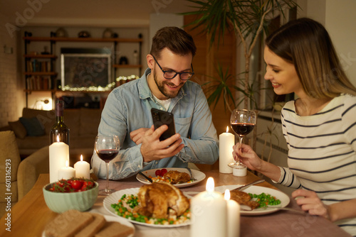 Happy couple enjoying food and wine while surfing the net on smart phone at home
