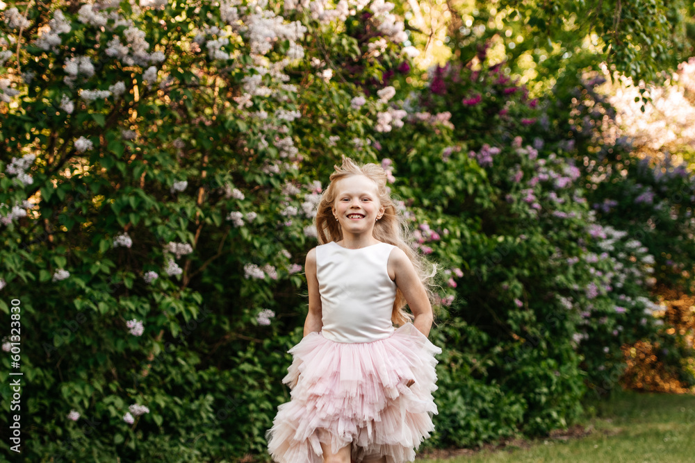 A beautiful blonde 8 years old runs in a festive dress in the park, against the backdrop of lilac bushes.
