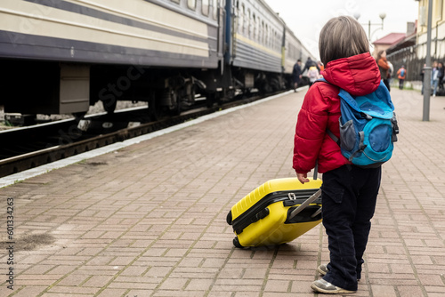 small baby boy stands on the platform near the train and holds a yellow suitcase in his hands.