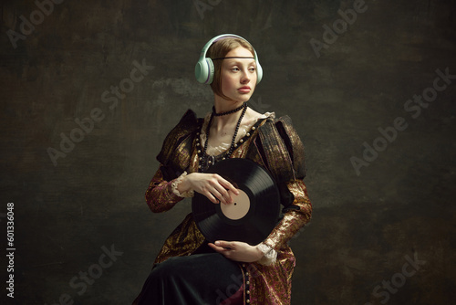 Portrait of pretty young girl, royal person in elegant vintage dress listening to music in headphones, holding vinyl on dark green background. Concept of history, renaissance art, comparison of eras
