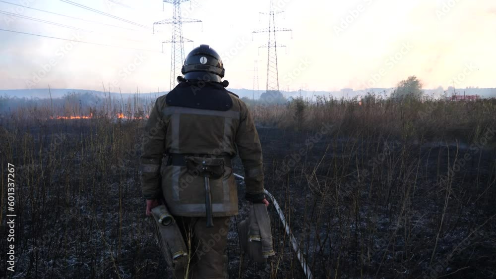 Male fireguard in full equipment walking with fire hoses on burnt grass at countryside. Young fireman in uniform going through charred meadow to extinguish fire. Concept of environmental issues Male fireguard in full equipment walking with fire hoses on burnt grass at countryside. Young fireman in uniform going through charred meadow to extinguish fire. Concept of environmental issues