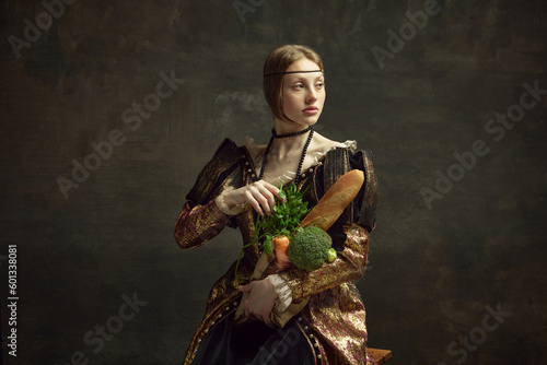 Portrait of young beautiful girl, princess in vintage, elegant costume with baguette and vegetables on dark green background. Concept of history, renaissance art, comparison of eras. Grocery shopping